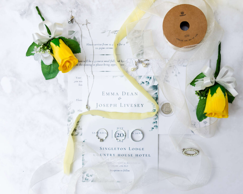 Wedding invitation flatlay featuring yellow rose buttonholes, white ribbons, jewellery, rings, a coin, and a spool arranged around an invitation card on a white marble surface. Copyright Somerside Photography Ltd // www.somersidephotography.com