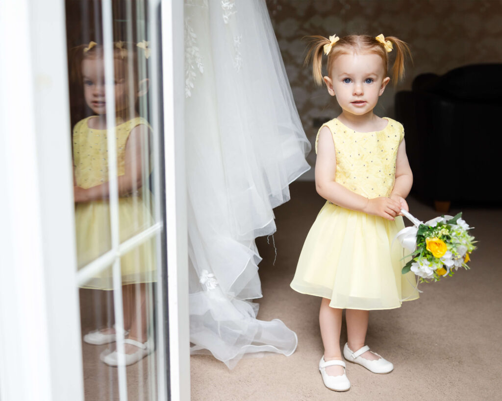 Young girl in a yellow dress holding a bunch of flowers stands indoors near a window, with her reflection visible on the glass. Copyright Somerside Photography Ltd // www.somersidephotography.com