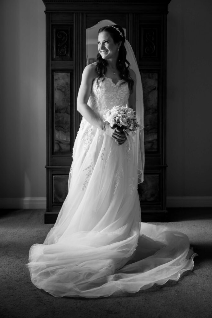 A bride in a lace wedding dress and veil stands indoors holding a bouquet, smiling and looking to the side. Copyright Somerside Photography Ltd // www.somersidephotography.com