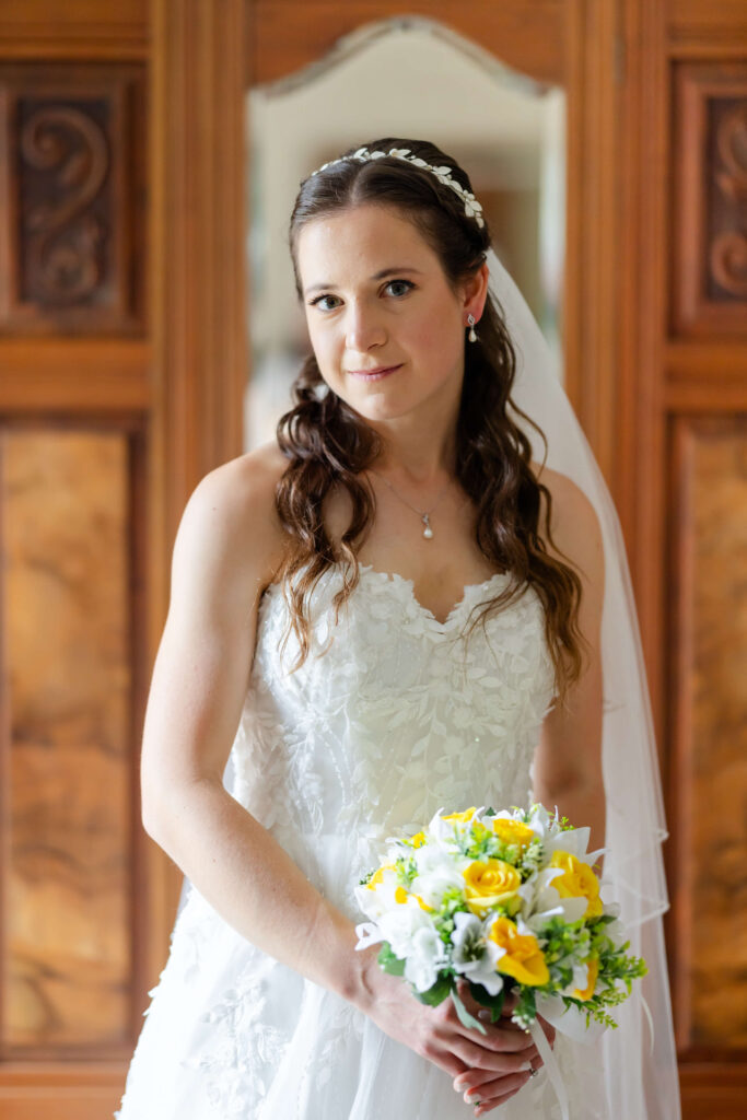 A woman in a white wedding dress holds a bouquet of yellow and white flowers, standing indoors in front of a wooden wardrobe with a mirror. Copyright Somerside Photography Ltd // www.somersidephotography.com