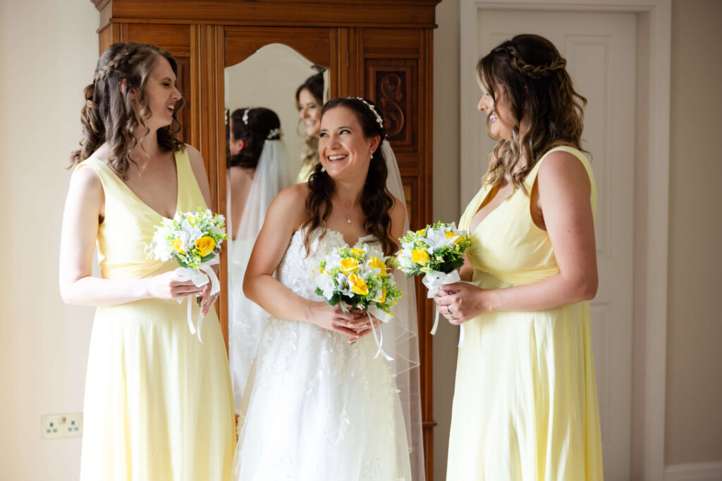 A bride in a white dress stands between two bridesmaids in yellow dresses, all holding bouquets and smiling indoors. Copyright Somerside Photography Ltd // www.somersidephotography.com