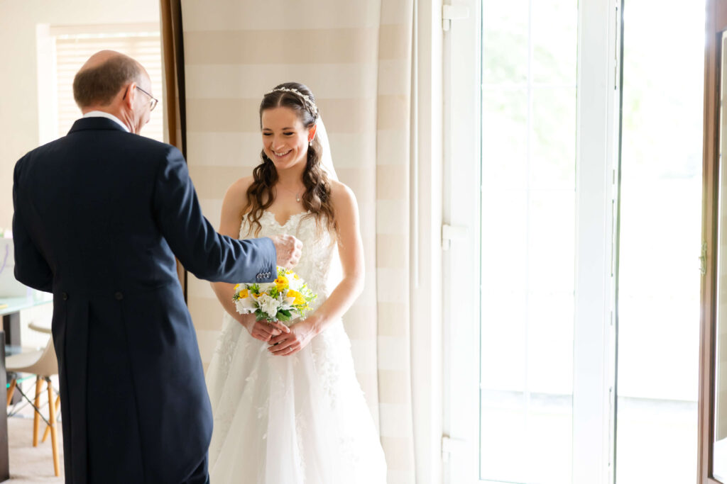 A bride in a white wedding dress holding a bunch of yellow flowers smiles at an older man in a suit indoors near a window. Copyright Somerside Photography Ltd // www.somersidephotography.com