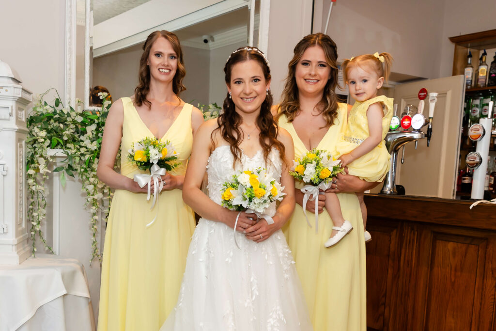 A bride in a white dress stands with two bridesmaids in yellow dresses and a young girl in yellow, all holding yellow and white bouquets, posing indoors beside a bar. Copyright Somerside Photography Ltd // www.somersidephotography.com