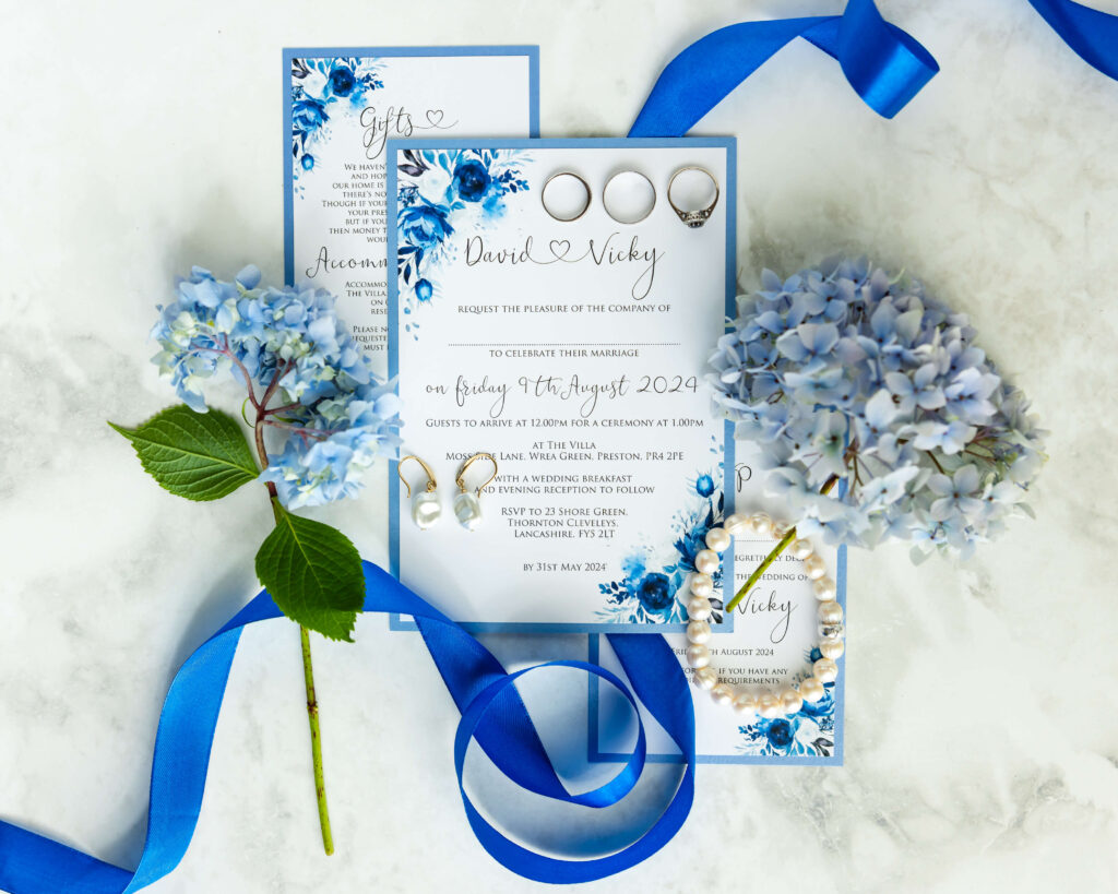 Flat lay of wedding invitations with blue floral designs, silver rings, pearl jewellery, blue ribbon, and light blue hydrangea flowers on a white surface. Copyright Somerside Photography Ltd // www.somersidephotography.com