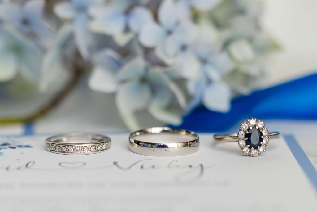 Three rings—a diamond band, a plain wedding band, and a sapphire halo ring—are displayed on a card with blurred blue and white flowers in the background. Copyright Somerside Photography Ltd // www.somersidephotography.com