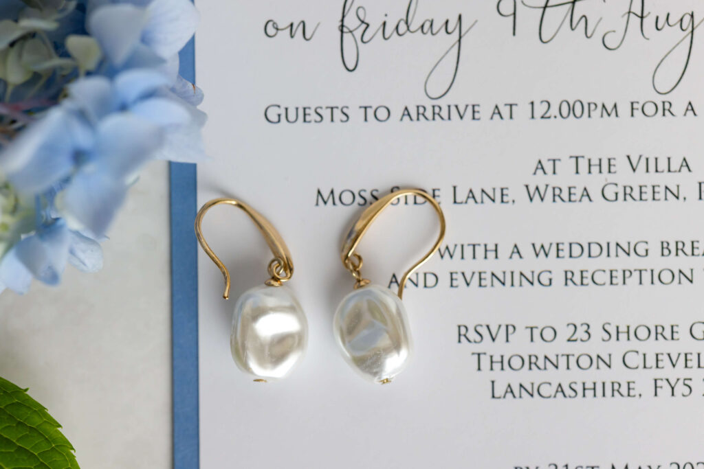 A pair of gold and pearl drop earrings placed on a wedding invitation card with floral decorations nearby. Copyright Somerside Photography Ltd // www.somersidephotography.com