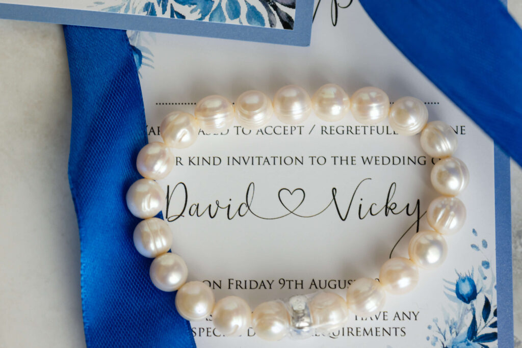 A pearl bracelet and blue ribbon rest on a wedding invitation card with the names David and Vicky written in joined-up writing. Copyright Somerside Photography Ltd // www.somersidephotography.com