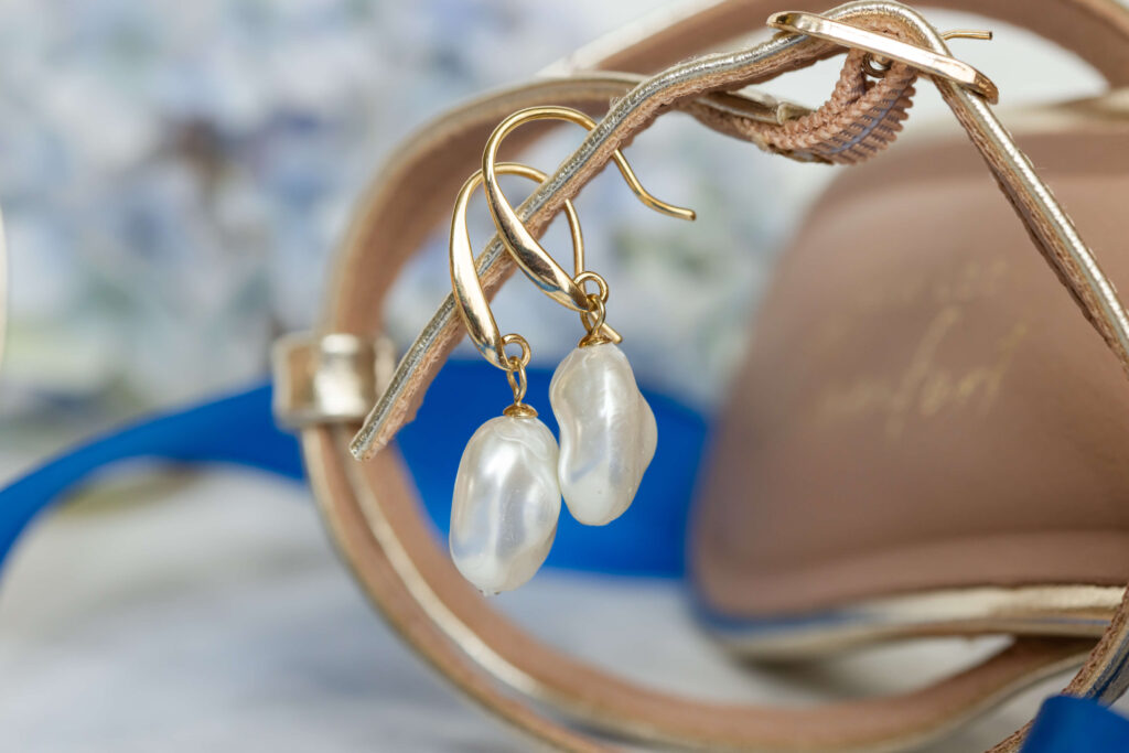 A pair of gold earrings with irregular white pearls hanging on the strap of a beige sandal. Copyright Somerside Photography Ltd // www.somersidephotography.com