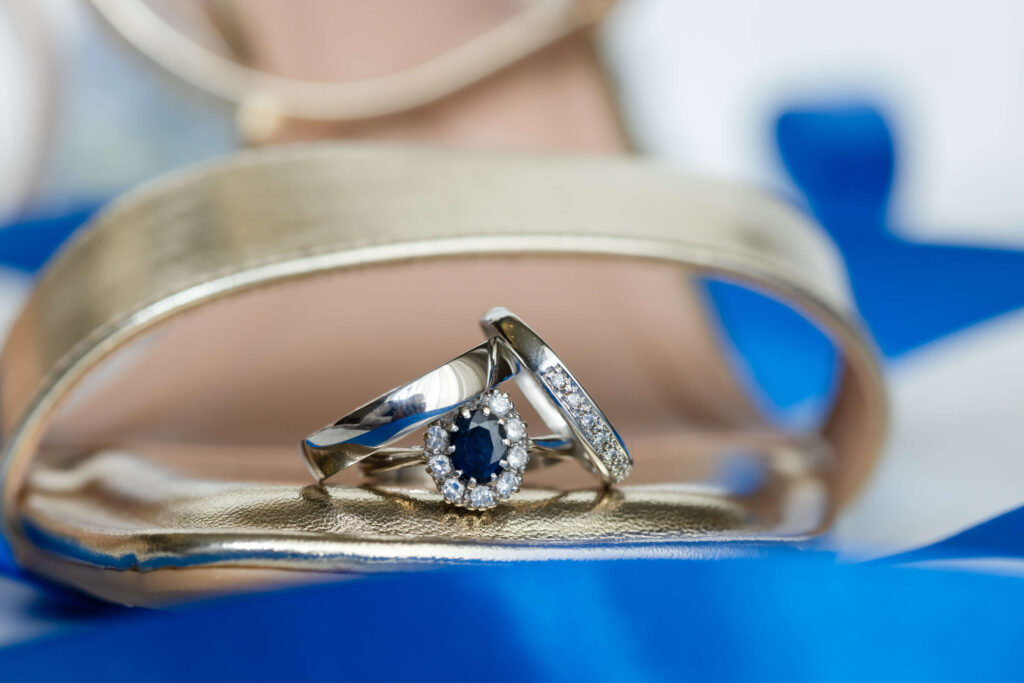 Close-up of a pair of wedding rings, one with a blue gemstone and diamonds, resting on a gold sandal with a blurred blue and white background. Copyright Somerside Photography Ltd // www.somersidephotography.com