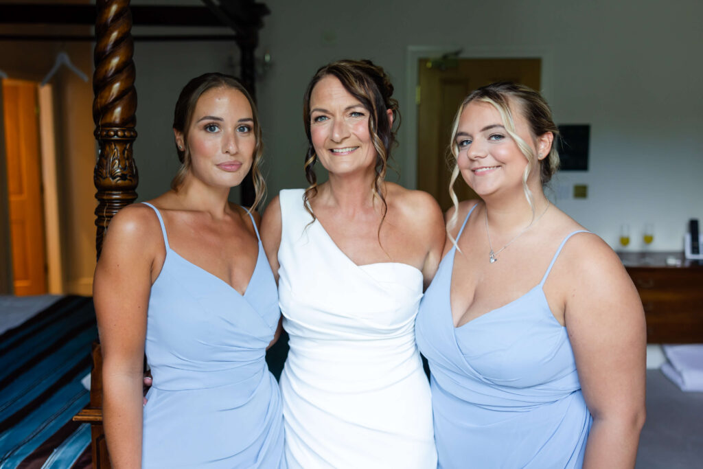 Three women pose indoors; the woman in the centre wears a white dress, while the two on either side wear matching light blue dresses. Copyright Somerside Photography Ltd // www.somersidephotography.com
