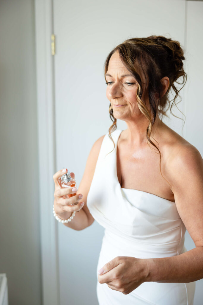 Woman in a white one-shoulder dress holds a small perfume bottle, preparing to spray it, standing indoors with a neutral background. Copyright Somerside Photography Ltd // www.somersidephotography.com