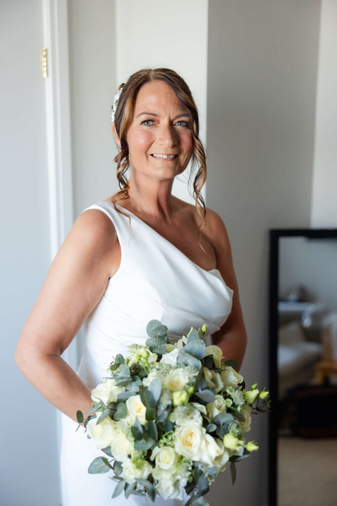 A woman in a white sleeveless dress holds a bunch of white and green flowers, smiling whilst posing indoors near a mirror. Copyright Somerside Photography Ltd // www.somersidephotography.com