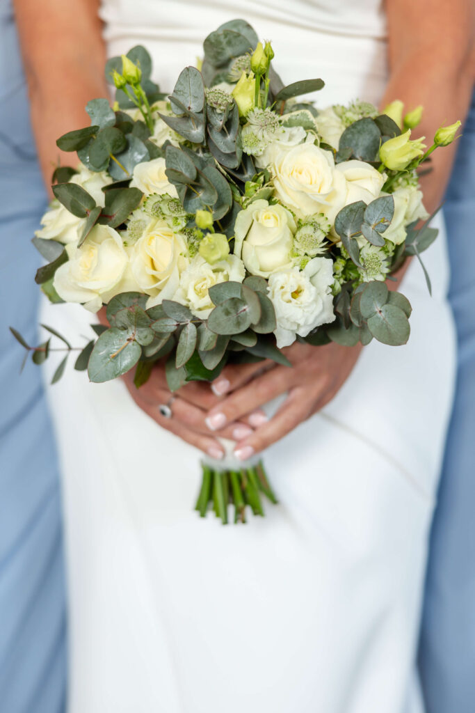 A person in a white dress holds a bouquet of white and pale yellow roses, greenery, and eucalyptus leaves. Copyright Somerside Photography Ltd // www.somersidephotography.com