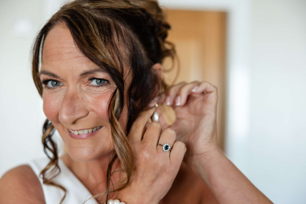 Woman with wavy brown hair smiles while putting on an earring, wearing a white top and a ring with a dark stone. Copyright Somerside Photography Ltd // www.somersidephotography.com