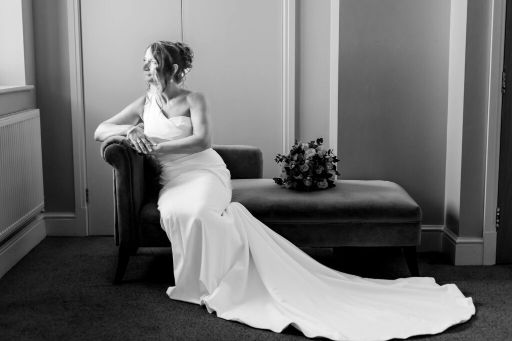 Black and white photo of a bride in a fitted wedding dress seated on a chaise longue, looking to one side, with a floral bouquet resting beside her. Copyright Somerside Photography Ltd // www.somersidephotography.com