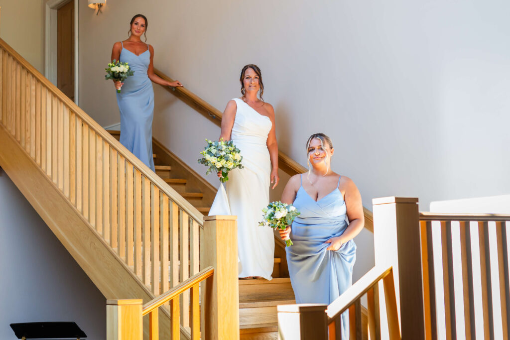 Three women in light blue and white dresses holding bouquets stand on a wooden staircase, posing for a photo in a brightly lit indoor setting. Copyright Somerside Photography Ltd // www.somersidephotography.com