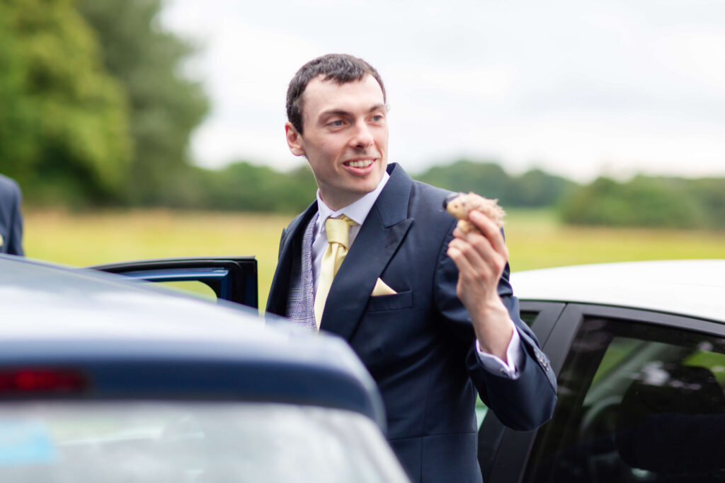 A man in a suit stands next to a car, holding a small object in his right hand, with greenery and trees in the background. Copyright Somerside Photography Ltd // www.somersidephotography.com