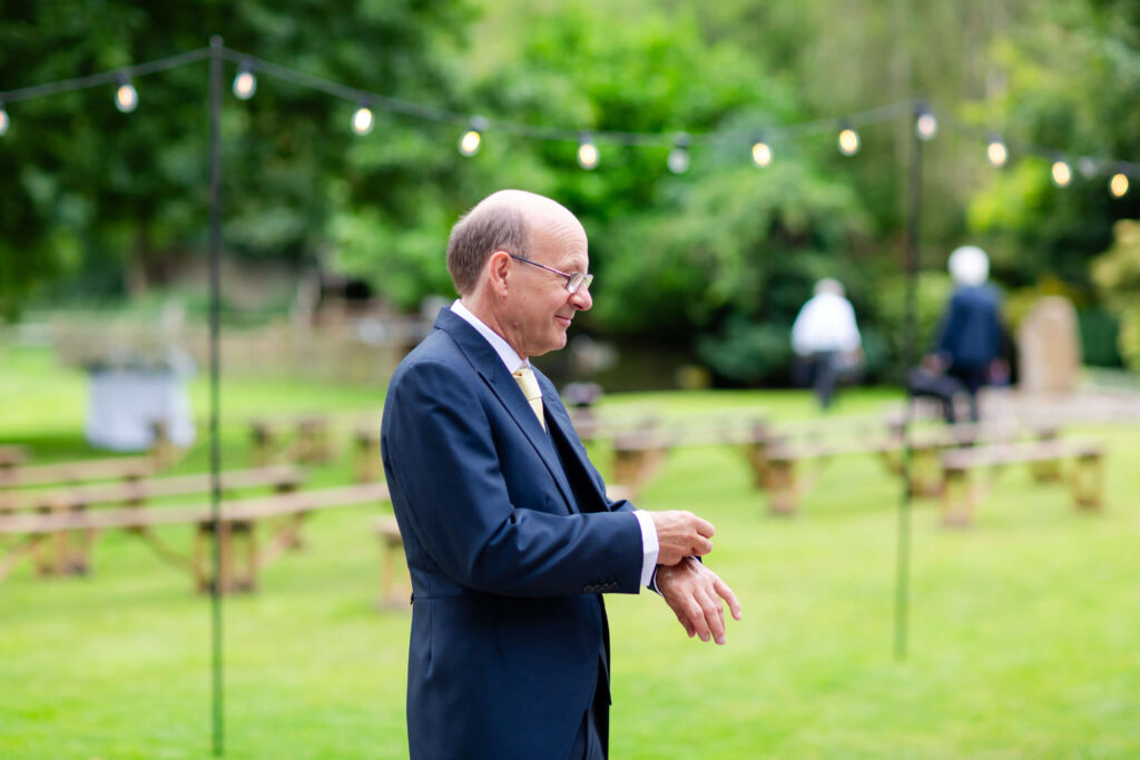 An older man in a suit stands on a grassy lawn with fairy lights and wooden benches in the background, looking to the side. Copyright Somerside Photography Ltd // www.somersidephotography.com