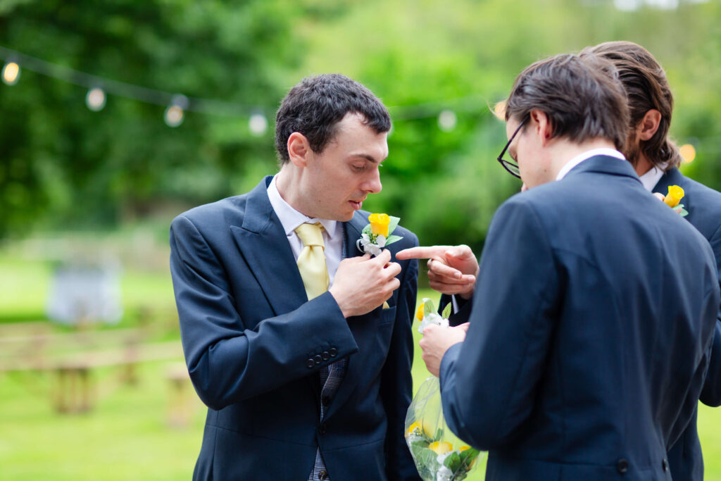 Three men in suits stand outdoors. One man adjusts a yellow buttonhole on his lapel while another points at it. The background is green and blurred. Copyright Somerside Photography Ltd // www.somersidephotography.com