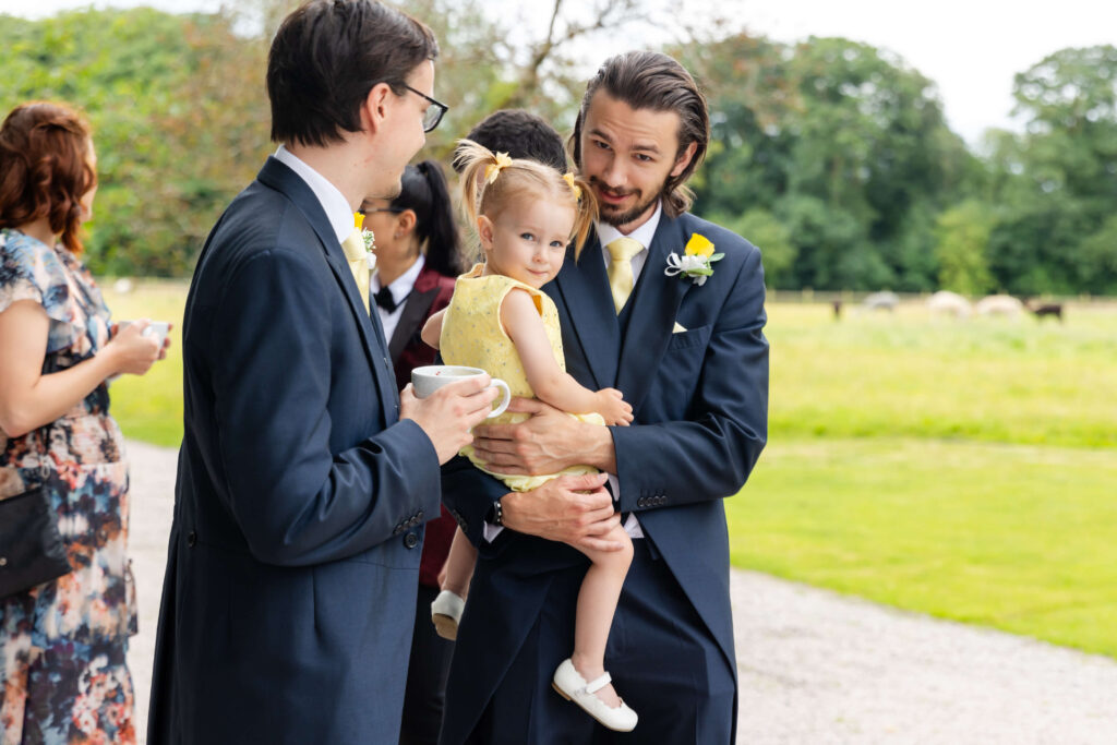 Two men in suits talk outside while one holds a young girl in a yellow dress. Other people and greenery are visible in the background. Copyright Somerside Photography Ltd // www.somersidephotography.com