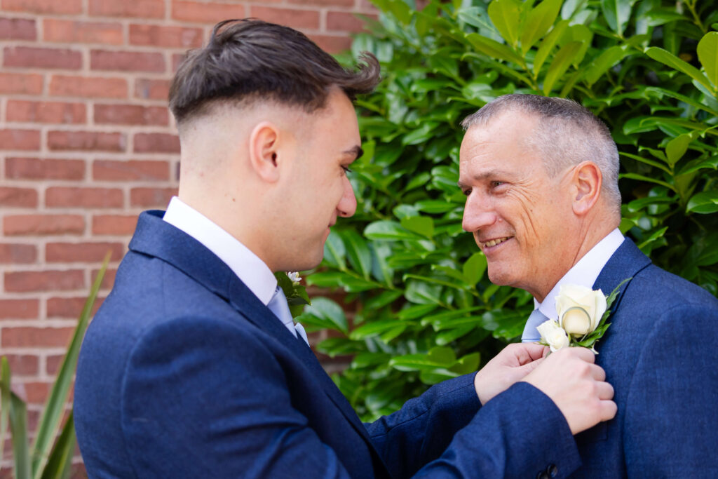 Two men in suits stand outside; one is pinning a white rose buttonhole onto the other's jacket. They are facing each other and smiling, with greenery and a brick wall in the background. Copyright Somerside Photography Ltd // www.somersidephotography.com