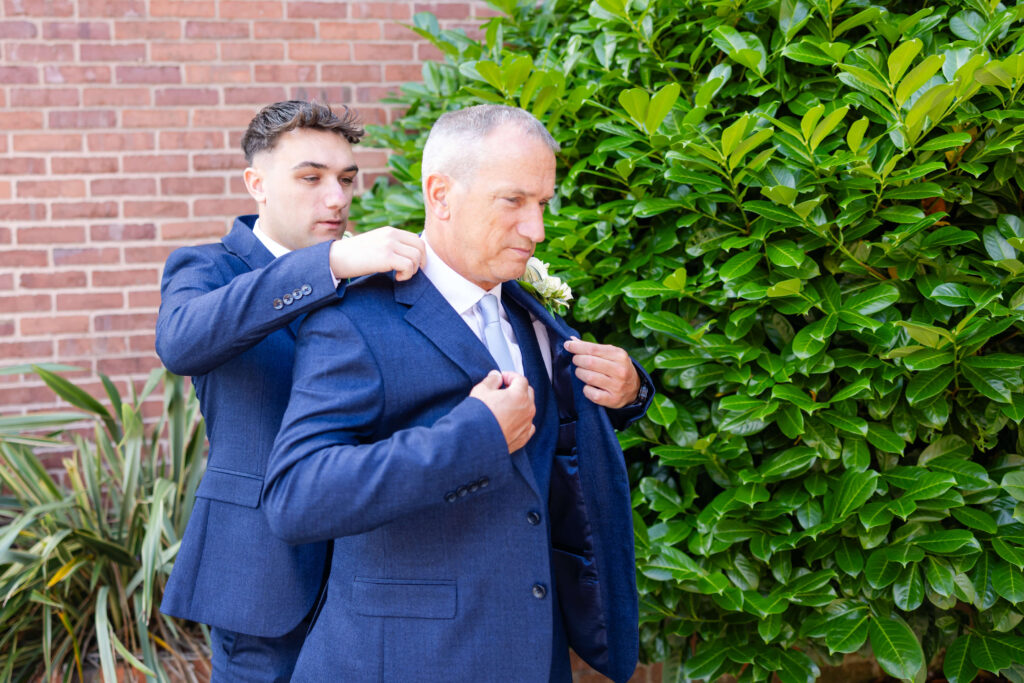 A young man helps an older man adjust his suit jacket outside near a brick wall and green bush. Copyright Somerside Photography Ltd // www.somersidephotography.com