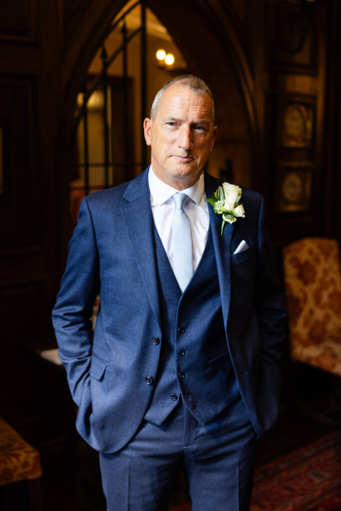 A man in a blue suit with a white shirt and tie stands indoors with hands in pockets, wearing a white rose buttonhole. Copyright Somerside Photography Ltd // www.somersidephotography.com