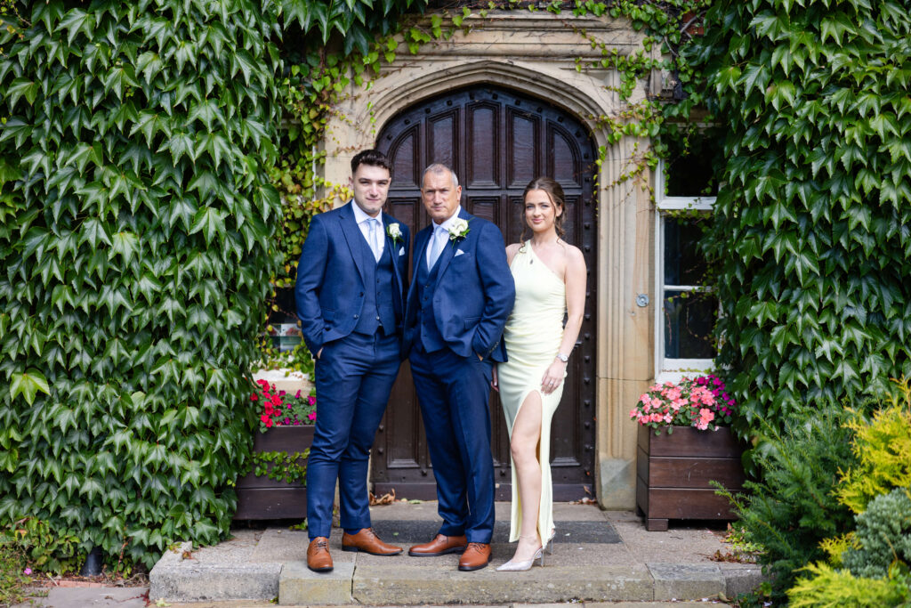 Three people dressed formally stand in front of an arched wooden doorway framed by ivy, with two planters of pink flowers on either side. Copyright Somerside Photography Ltd // www.somersidephotography.com