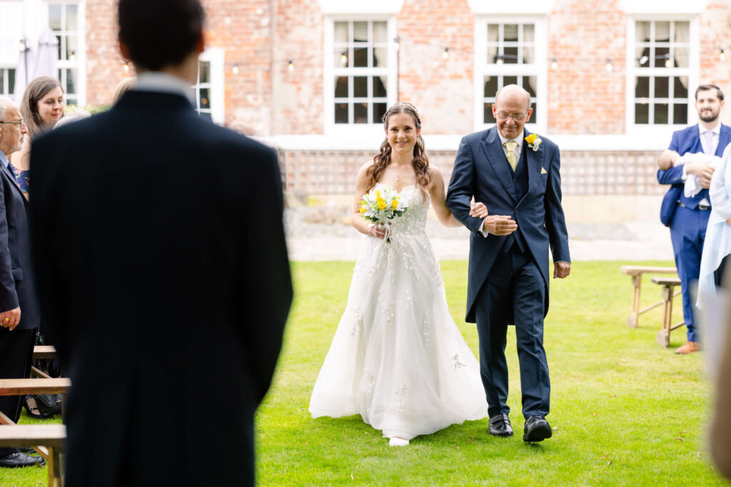 A bride in a white dress walks down the aisle on green grass, holding a bouquet and accompanied by an older man in a suit, with guests seated on either side. Copyright Somerside Photography Ltd // www.somersidephotography.com