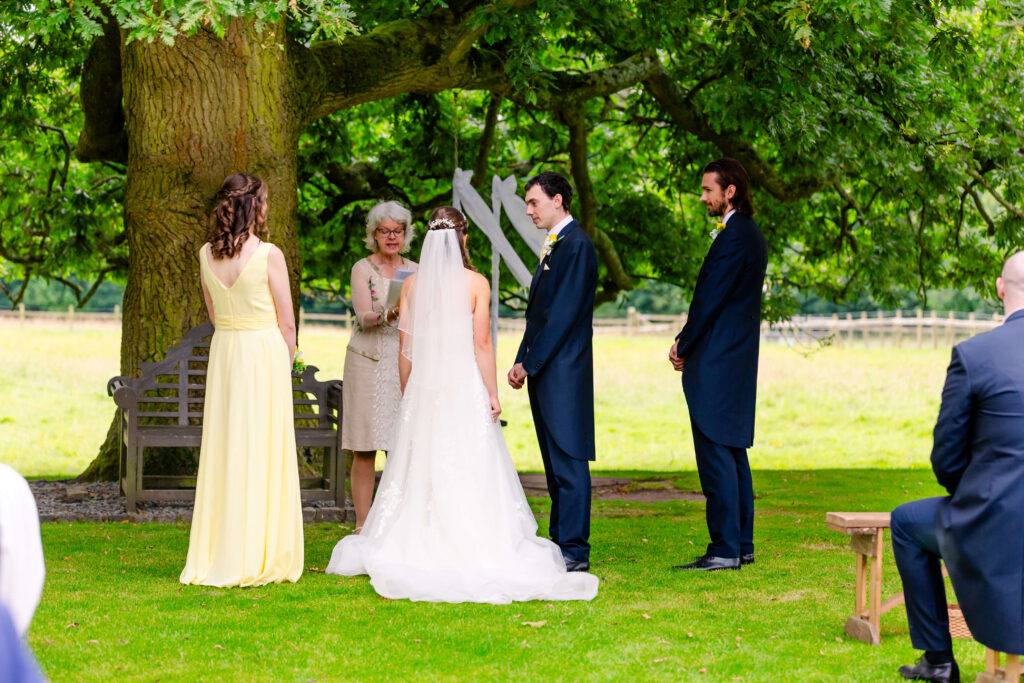 An outdoor wedding ceremony under a large tree, with a bride and groom standing before an officiant, accompanied by a bridesmaid and a best man. Copyright Somerside Photography Ltd // www.somersidephotography.com