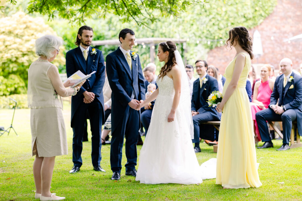 A couple stands holding hands during an outdoor wedding ceremony, with an officiant reading and guests seated in the background. Copyright Somerside Photography Ltd // www.somersidephotography.com