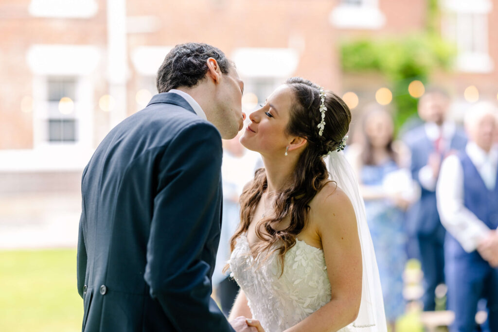 A bride and groom kiss outdoors during their wedding ceremony, with blurred guests and greenery in the background. Copyright Somerside Photography Ltd // www.somersidephotography.com