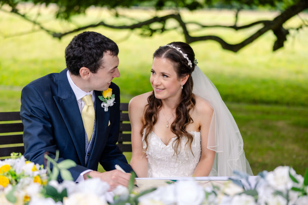 A bride and groom sit together at a table outdoors, looking at each other and smiling, with flowers and greenery in the foreground. Copyright Somerside Photography Ltd // www.somersidephotography.com