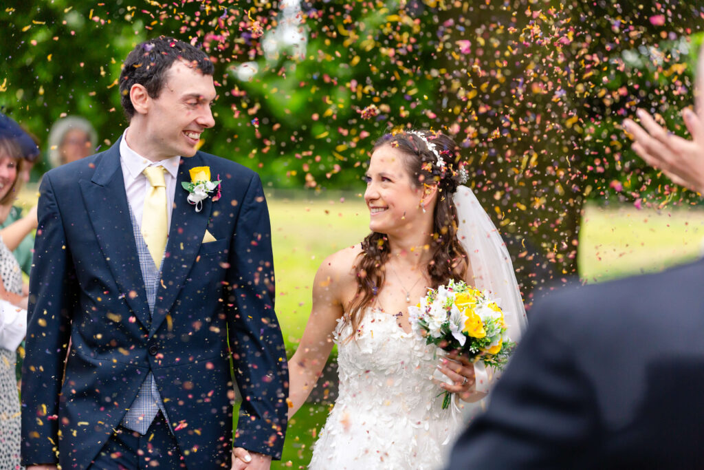 A bride and groom walk outdoors, smiling at each other, as confetti is thrown around them. The bride holds a bouquet and wears a white dress; the groom is in a dark suit. Copyright Somerside Photography Ltd // www.somersidephotography.com