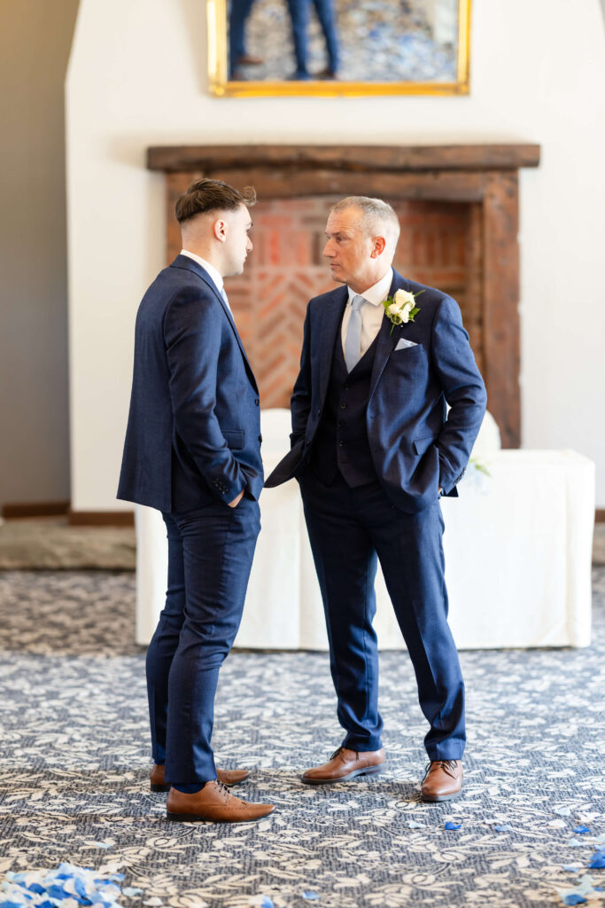 Two men in suits stand facing each other and talking in a formal setting with a fireplace and mirror in the background. Copyright Somerside Photography Ltd // www.somersidephotography.com