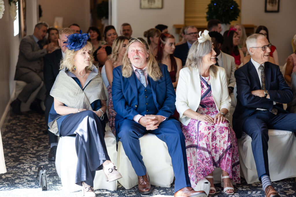 A group of people in formal attire sit and smile in rows of chairs at an indoor event, with sunlight streaming through a window. Copyright Somerside Photography Ltd // www.somersidephotography.com