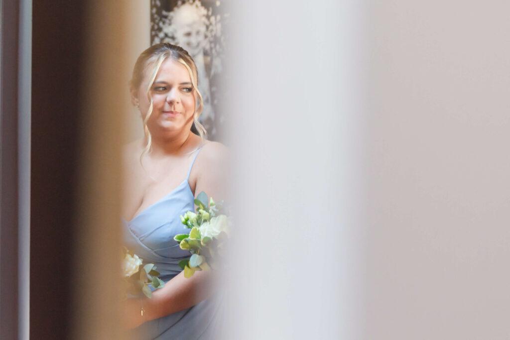 A woman in a light blue dress holding a bunch of flowers stands indoors, partially obscured by a blurred foreground. Copyright Somerside Photography Ltd // www.somersidephotography.com