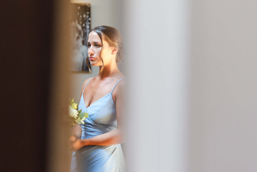 A woman in a light blue dress holds a small posy, standing indoors and looking to the side, partially framed by blurred white foreground. Copyright Somerside Photography Ltd // www.somersidephotography.com
