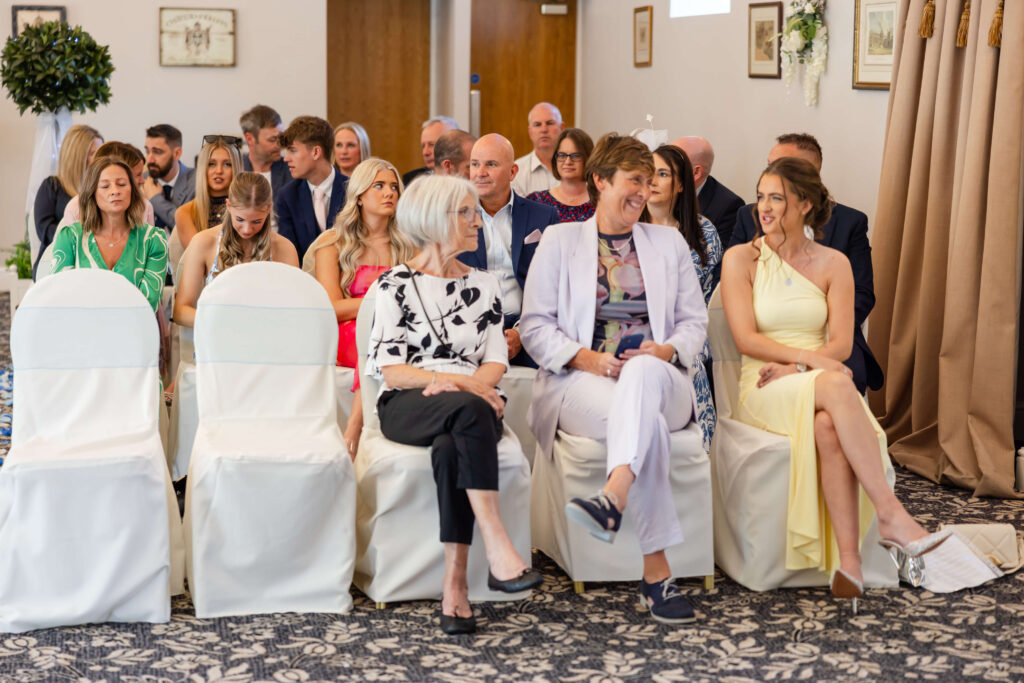 A group of people, both seated and standing, gather in a decorated indoor venue, suggesting a formal event or ceremony. Three women sit in the front row, conversing. Copyright Somerside Photography Ltd // www.somersidephotography.com