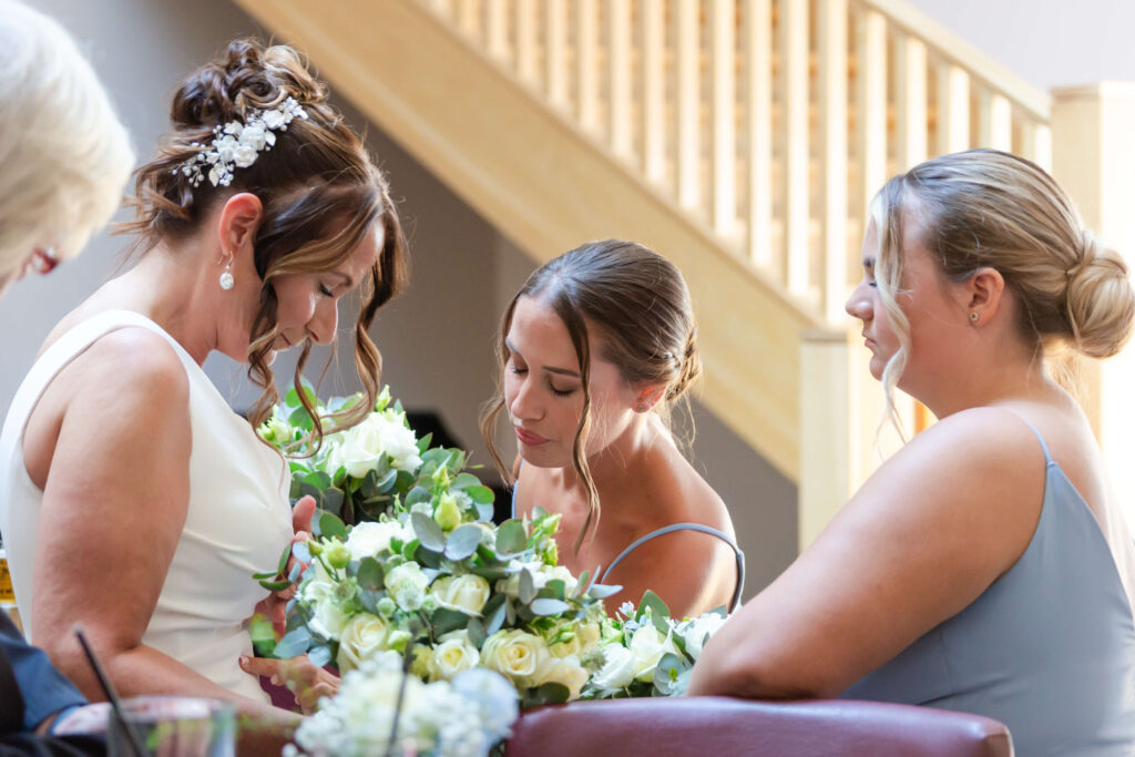 Three women in formal dresses sit closely together indoors, holding bouquets of white and green flowers, with a wooden staircase in the background. Copyright Somerside Photography Ltd // www.somersidephotography.com