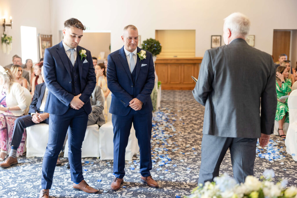 Two men in blue suits stand at the front of a room during a wedding ceremony, facing an officiant. Guests are seated on white chairs, some watching the proceedings. Copyright Somerside Photography Ltd // www.somersidephotography.com