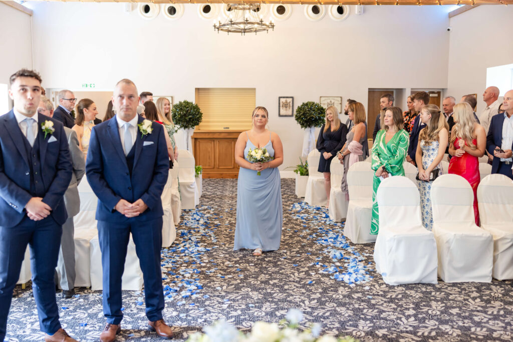 A bridesmaid walks down an aisle lined with blue flower petals, whilst guests and two men in suits stand at the front in a decorated indoor venue. Copyright Somerside Photography Ltd // www.somersidephotography.com