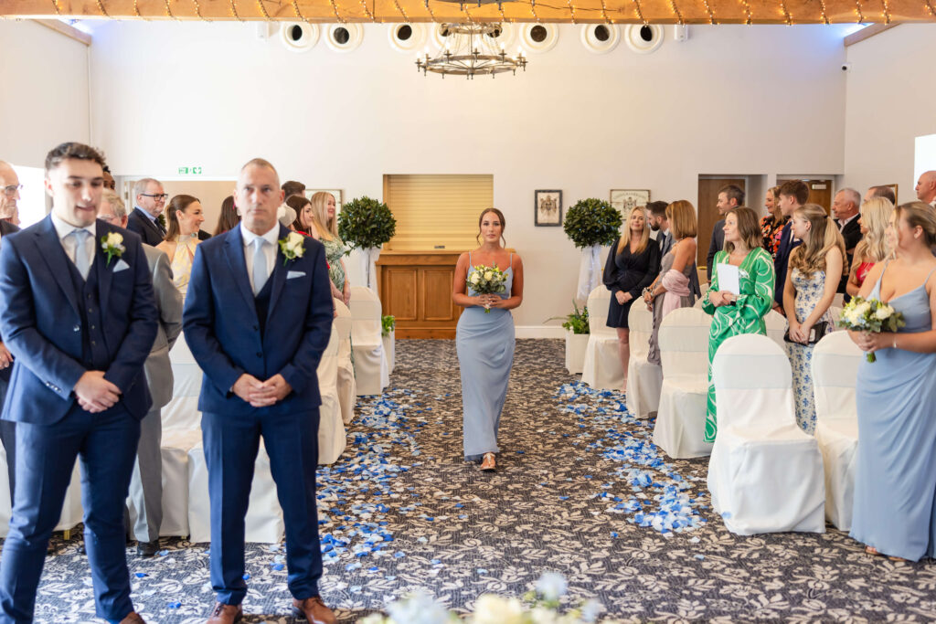 A bridesmaid walks down the aisle holding a bouquet whilst guests seated on either side watch during a wedding ceremony in a decorated indoor venue. Copyright Somerside Photography Ltd // www.somersidephotography.com