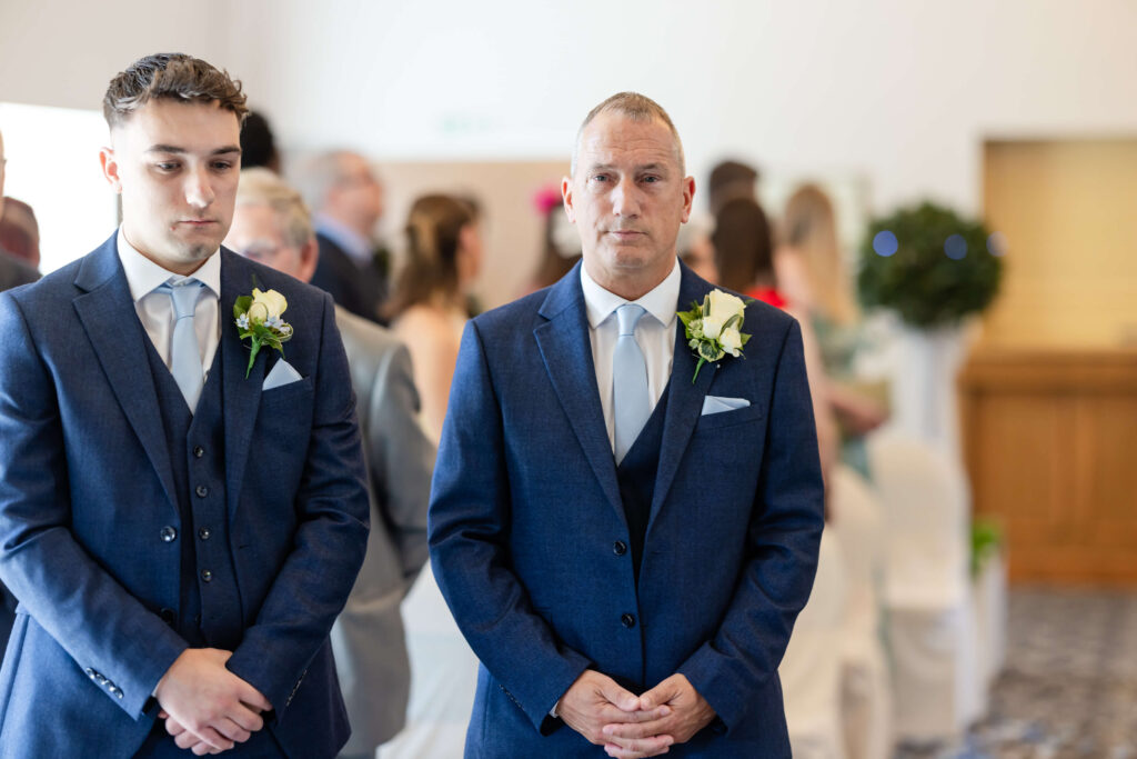 Two men in blue suits with white rose buttonholes stand indoors at a formal event, with blurred attendees seated in the background. Copyright Somerside Photography Ltd // www.somersidephotography.com