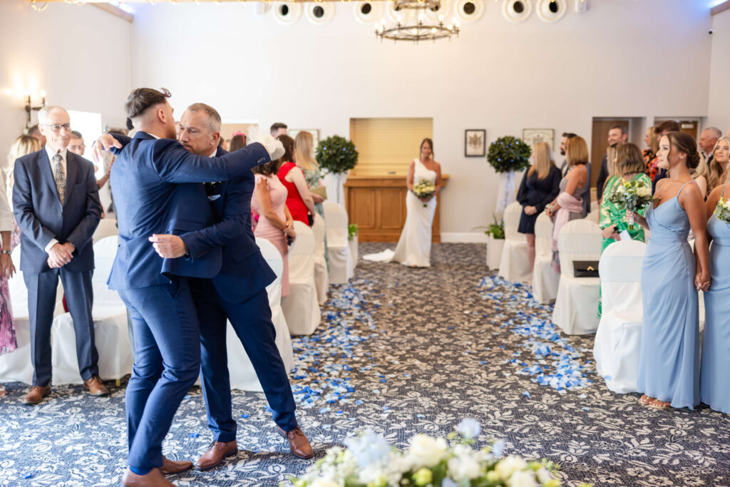 Two men in navy suits embrace in a wedding ceremony room as guests look on. A bride in white stands at the altar, with bridesmaids in blue nearby. Blue petals line the aisle. Copyright Somerside Photography Ltd // www.somersidephotography.com