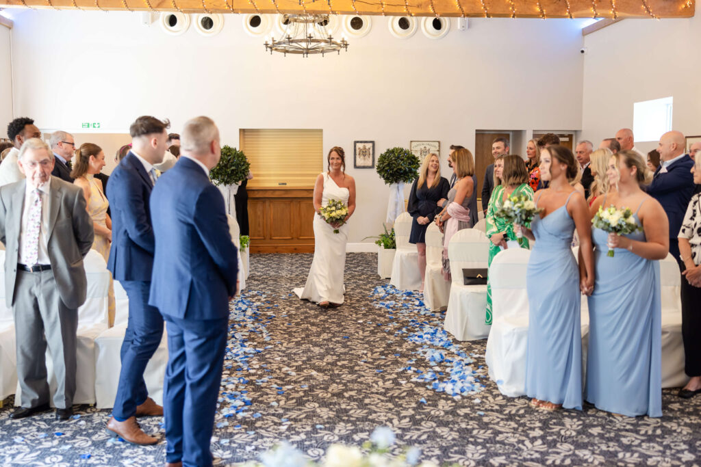 A bride in a white dress walks down the aisle with guests standing on both sides and bridesmaids in light blue dresses holding bouquets. Copyright Somerside Photography Ltd // www.somersidephotography.com
