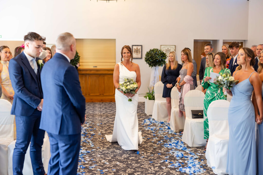 A bride in a white dress walks down the aisle holding a bouquet, approaching a groom as guests stand on either side in a decorated indoor venue. Copyright Somerside Photography Ltd // www.somersidephotography.com