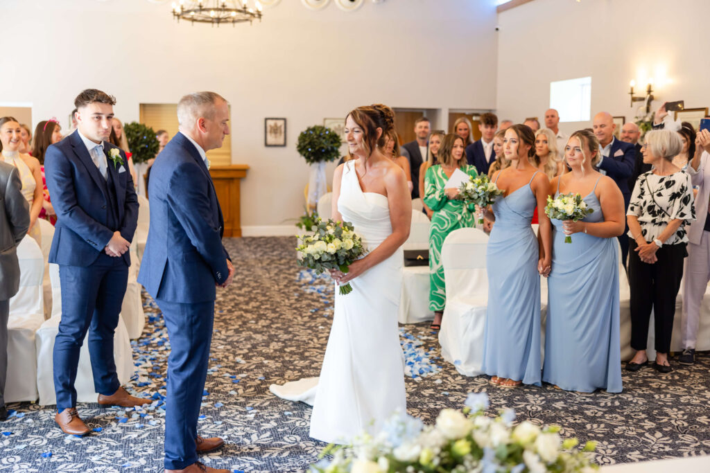 A bride in a white dress stands with her bouquet, facing a man in a suit, surrounded by bridesmaids in blue dresses and guests at an indoor wedding ceremony. Copyright Somerside Photography Ltd // www.somersidephotography.com