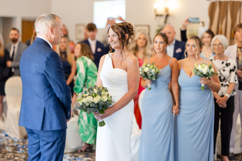 A bride in a white dress and a groom in a blue suit stand facing each other at a wedding ceremony, with bridesmaids and guests in the background. Copyright Somerside Photography Ltd // www.somersidephotography.com