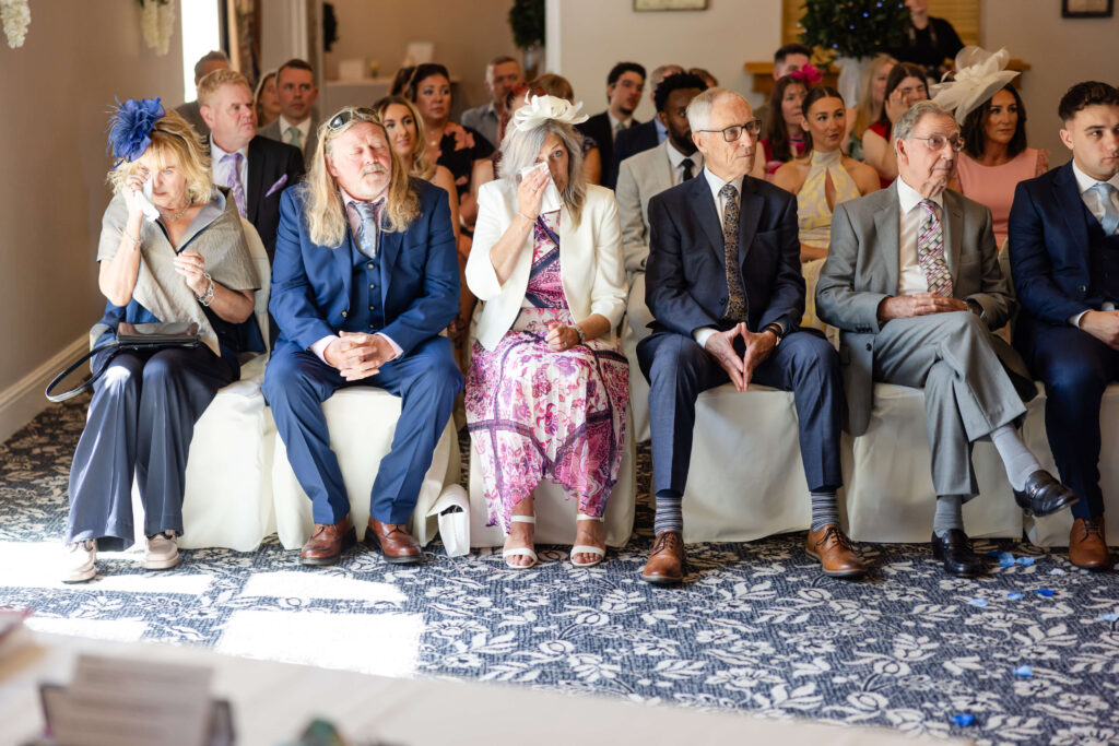 Group of formally dressed people seated in rows at an indoor event, likely a wedding ceremony, with some individuals wearing hats and fascinators. Copyright Somerside Photography Ltd // www.somersidephotography.com
